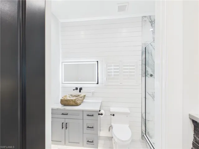Bathroom featuring a unique stone vessel sink, gray vanity with dark hardware, illuminated mirror, and a glass-enclosed shower with marble-look wall tile