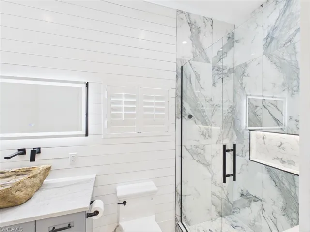 Bathroom featuring shiplap wall paneling, a vessel sink with a wall-mounted faucet, a vanity with a stone countertop, a glass-enclosed shower with marbled tile, and an illuminated shower niche