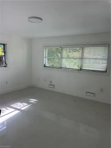 Room featuring a large window with horizontal blinds, a smaller window, a ceiling-mounted light fixture, white walls, and large format light-toned tile flooring