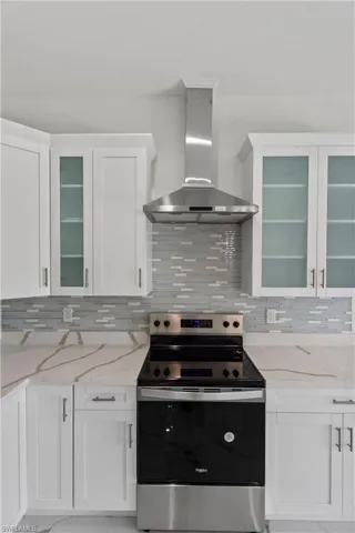 Kitchen featuring stainless steel electric range, wall chimney exhaust hood, tasteful backsplash, light marble finish floors, and white cabinetry