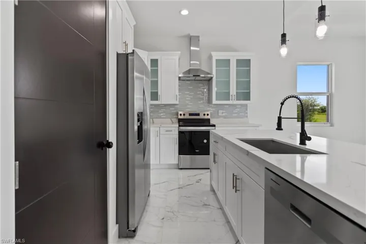 Kitchen featuring stainless steel appliances, decorative backsplash, wall chimney exhaust hood, light marble finish floors, and white cabinets