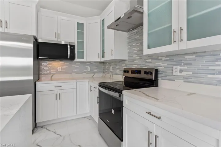 Kitchen featuring appliances with stainless steel finishes, decorative backsplash, light marble finish floors, wall chimney exhaust hood, and light stone countertops
