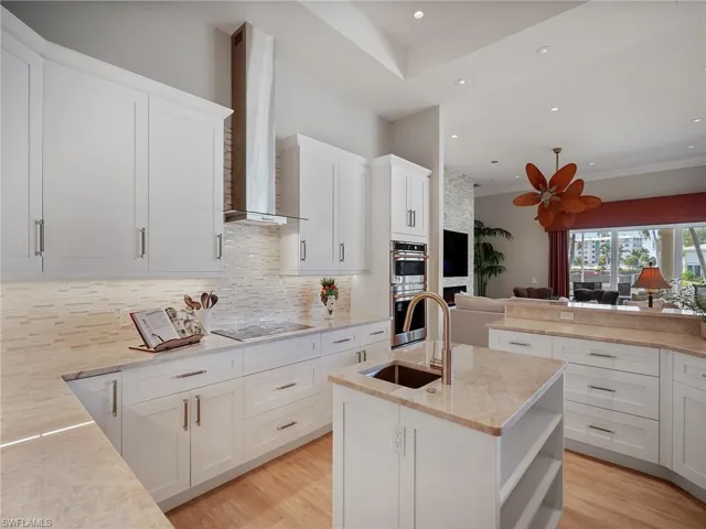 Kitchen featuring open shelves, a center island with sink, decorative backsplash, wall chimney range hood, and light stone countertops