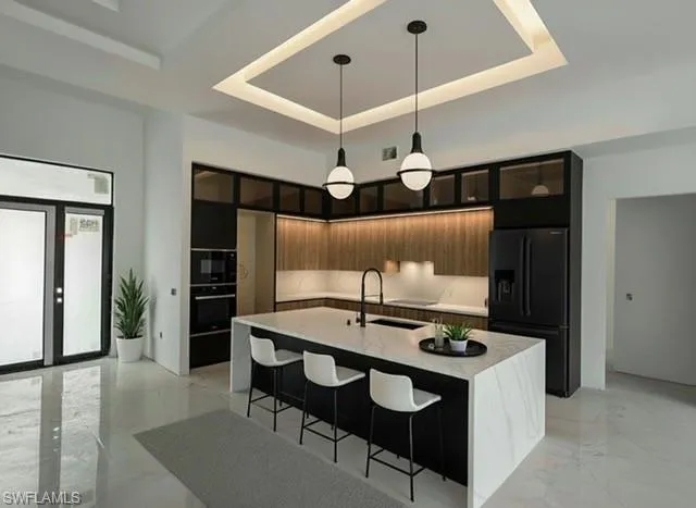 Kitchen featuring black refrigerator with ice dispenser, decorative light fixtures, an island with sink, modern cabinets, and light stone counters