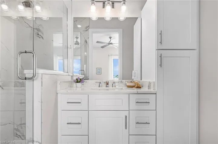 Bathroom featuring a single vanity with a white countertop and undermount sink, shaker-style cabinetry with chrome hardware, and a glass-enclosed shower with tile surround - Virtually Edited Image