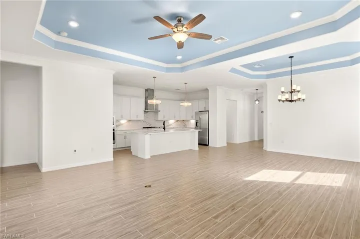 Unfurnished living room featuring a raised ceiling, a chandelier, ceiling fan, light wood-type flooring, and ornamental molding