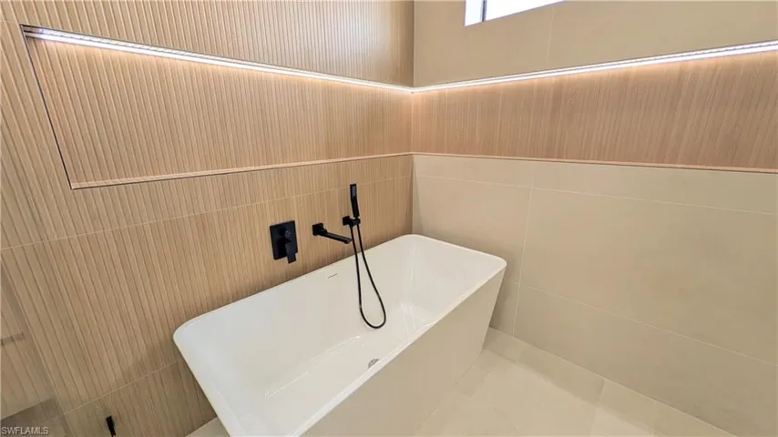 Freestanding white soaking tub with matte black wall-mounted faucet and handheld shower fixture, accented by integrated linear LED lighting and fluted wood-finish wall paneling