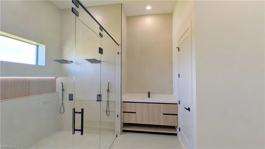 Walk-in glass-enclosed shower with dual rainfall showerheads, matte black fixtures, floating wood-finish vanity with light-toned countertop, and full-height wall tiling