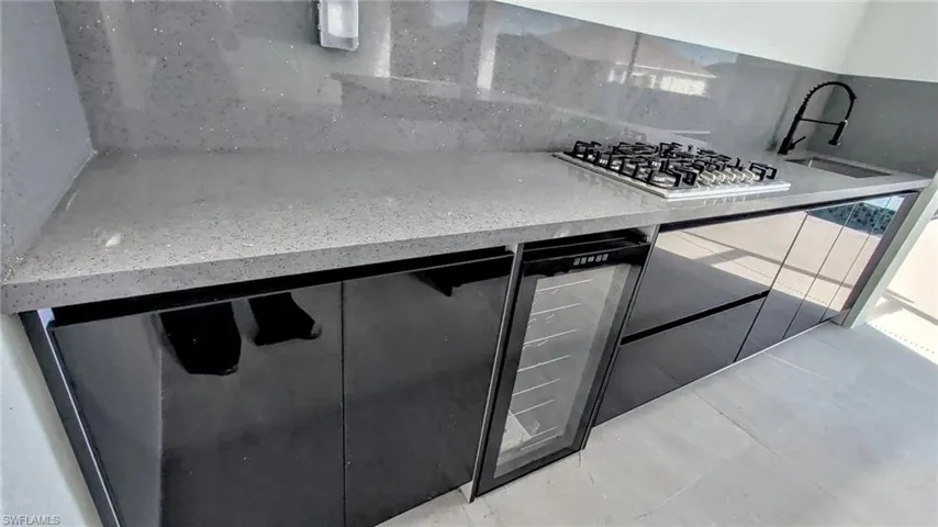 Outdoor kitchen featuring high-gloss black cabinetry, grey stone-finish countertops, an integrated gas cooktop, a built-in beverage refrigerator, and a gooseneck faucet