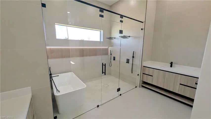 Wet room featuring a freestanding soaking tub, dual-head rainfall shower, floor-to-ceiling glass enclosure, wall-mounted matte black fixtures, and a floating vanity with wood-finish cabinetry