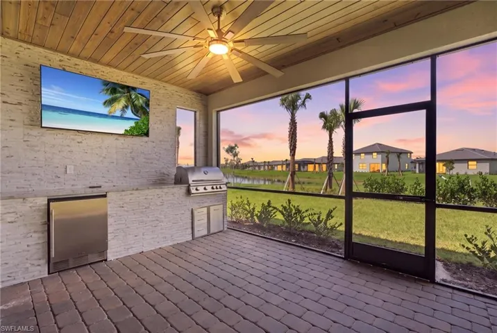Screened outdoor living area with a wood-plank ceiling and ceiling fan, featuring a built-in outdoor kitchen with a stainless steel grill and refrigeration unit, and paver flooring