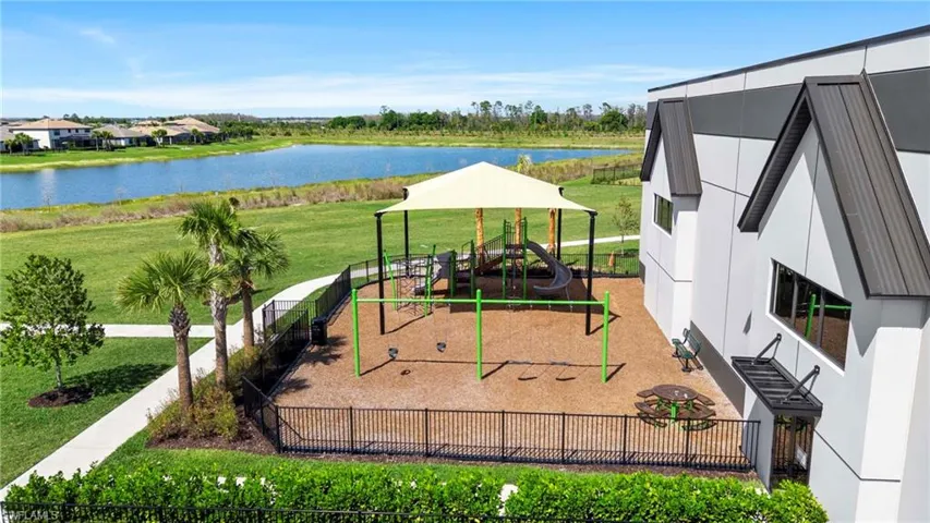 Community playground with shade canopy, swings, slides, and climbing structures, set beside a water feature, with surrounding manicured lawns and mature palm trees