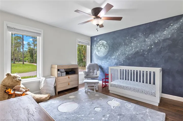 Bedroom with a crib, dark wood-style flooring, a ceiling fan, an accent wall, and wallpapered walls