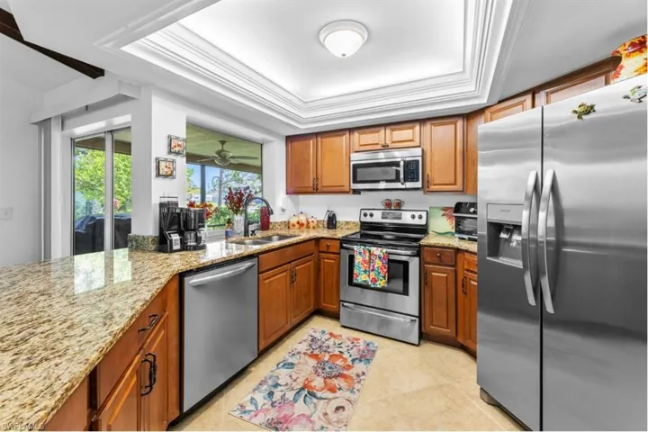 Kitchen featuring appliances with stainless steel finishes, a raised ceiling, brown cabinetry, light stone countertops, and ornamental molding
