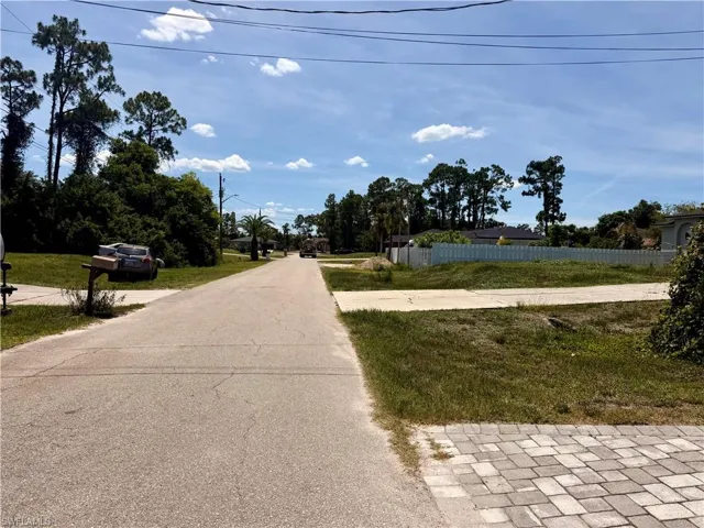 Residential street featuring paved road, mature trees, and open lot with partial perimeter fencing