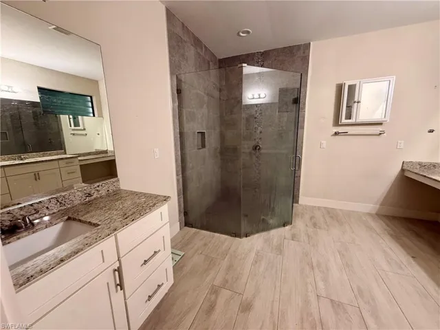 Spacious bathroom featuring a glass-enclosed shower with tile surround, dual vanity with undermount sinks, stone countertops, and wood-finish flooring