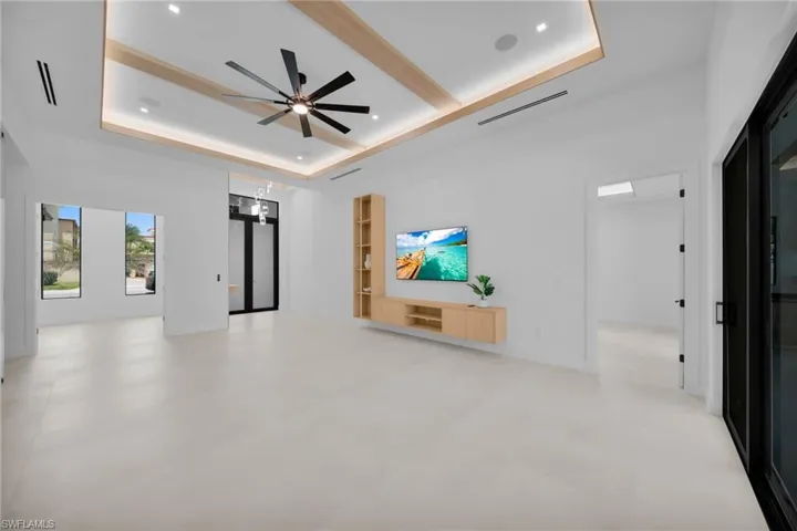 Spacious room featuring light-toned flooring, a tray ceiling with integrated lighting and wood-finish accents, and a contemporary ceiling fan