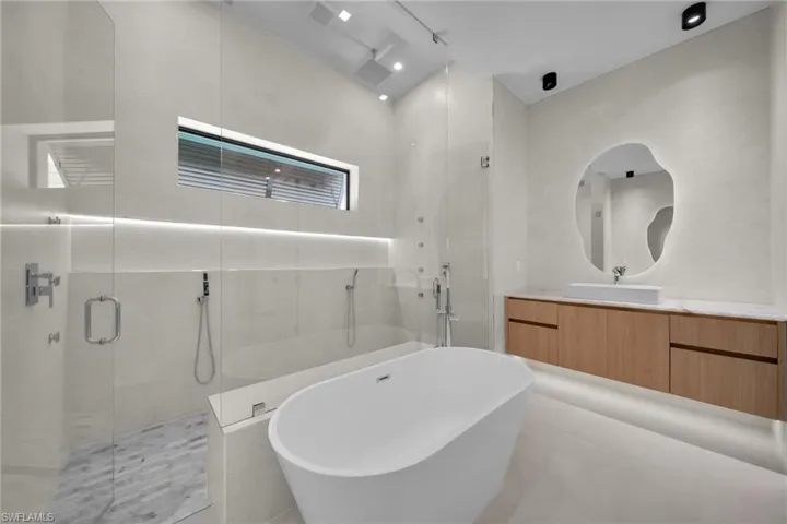 Contemporary bathroom featuring a freestanding soaking tub, a frameless glass shower enclosure with a tiled floor, a floating wood-finish vanity with a vessel sink, and an illuminated organic-shaped mirror