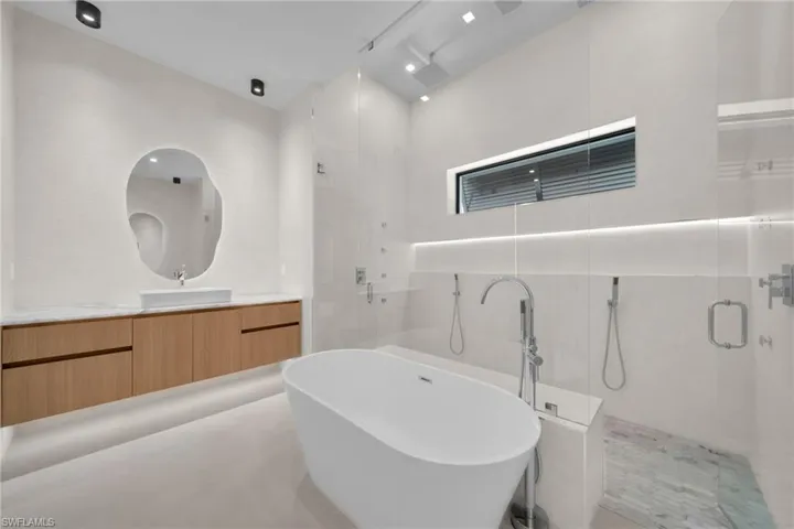Freestanding soaking tub with polished chrome floor-mounted filler, large walk-in shower with glass enclosure, floating wood-finish vanity with stone countertop and vessel sink, and a unique frameless mirror