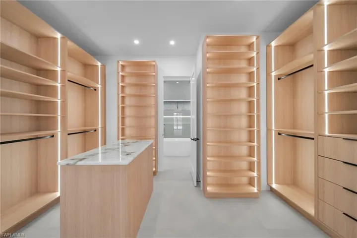 Custom closet system featuring light wood-finish cabinetry, integrated strip lighting, an island with a light-toned stone countertop, multiple shelving units, and hanging rods