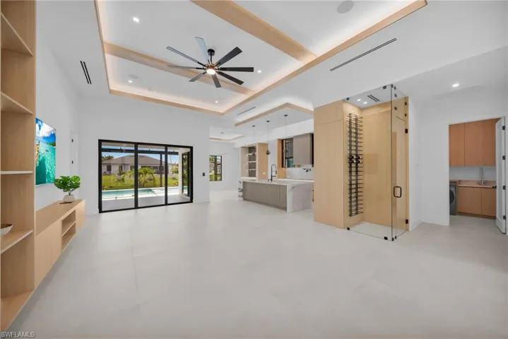 Spacious open-concept living area with light-toned tile flooring, a multi-paneled glass sliding door opening to an outdoor pool, a built-in media console with shelving, and a glass-enclosed wine display