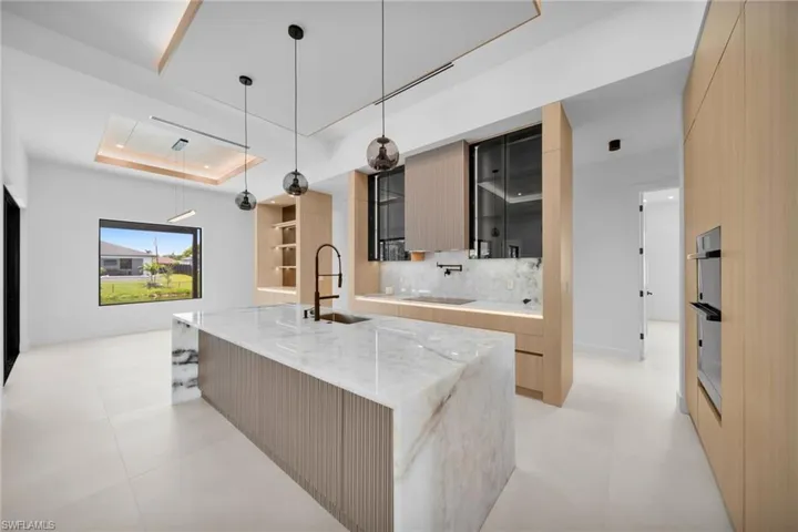 Gourmet kitchen with a waterfall edge island, integrated sink with a matte black faucet, light wood-finish cabinetry, built-in wall ovens, and contemporary pendant lighting