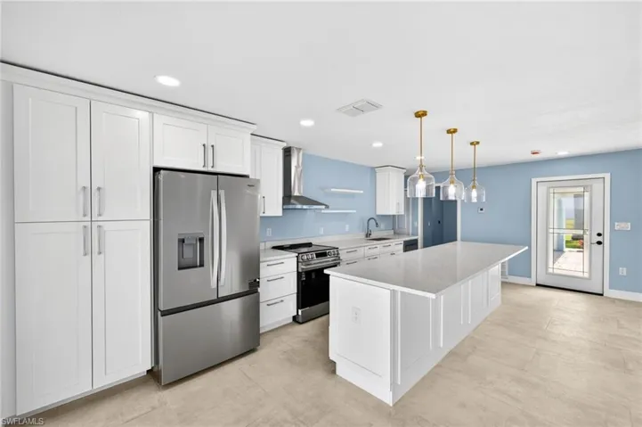 Modern kitchen featuring white shaker cabinetry, stainless steel appliances, a center island with a light-toned countertop, three glass pendant lights with brass accents, and light-toned tile flooring