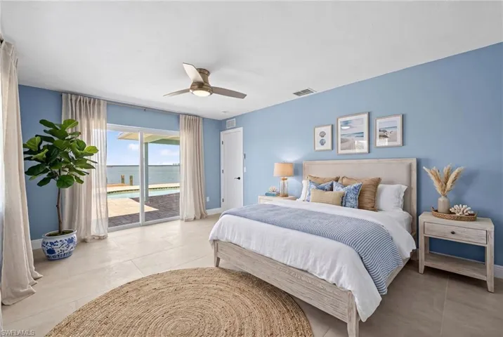 Virtually Staged ... Master bedroom featuring light blue walls, sliding glass doors opening to a waterfront, a ceiling fan, and light-toned tile flooring
