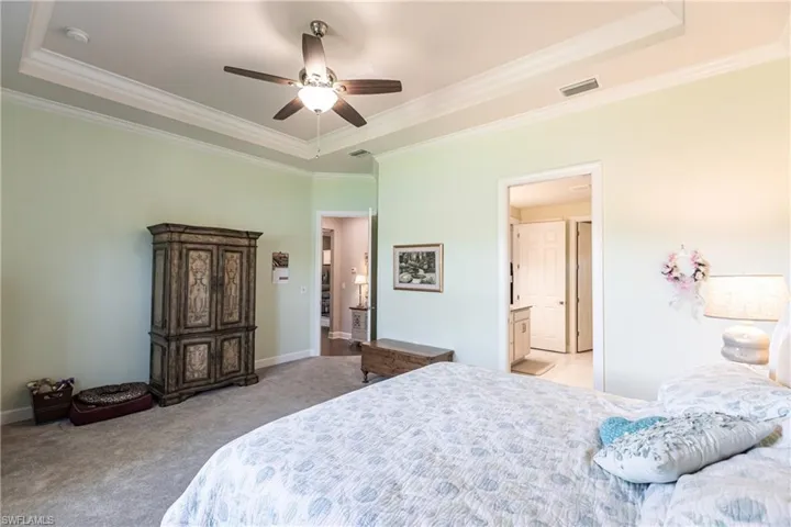 Carpeted bedroom featuring ceiling fan, ensuite bath, a tray ceiling, and crown molding