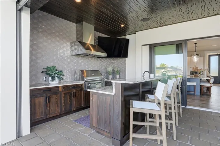 Outdoor kitchen with light stone countertops, a kitchen breakfast bar, wall chimney range hood, decorative backsplash, and a kitchen island with sink