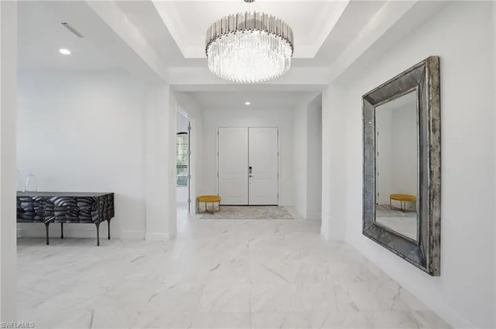 Entry foyer featuring double white paneled doors, tray ceiling with a large crystal chandelier, and white marble-look tile flooring
