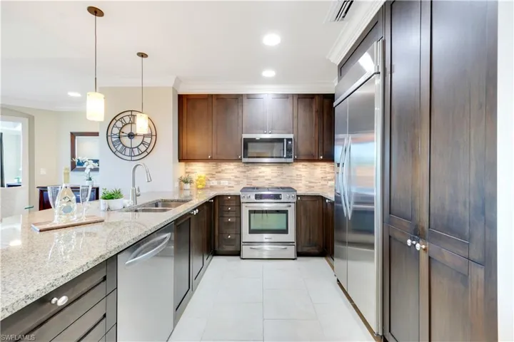 Kitchen with stainless steel appliances, dark brown cabinetry, backsplash, ornamental molding, and light stone counters