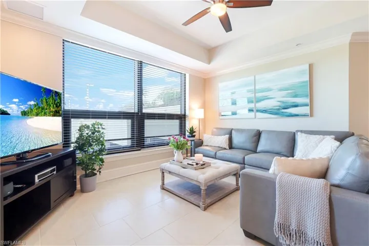 Living room with a raised ceiling, ornamental molding, light tile patterned floors, and a ceiling fan