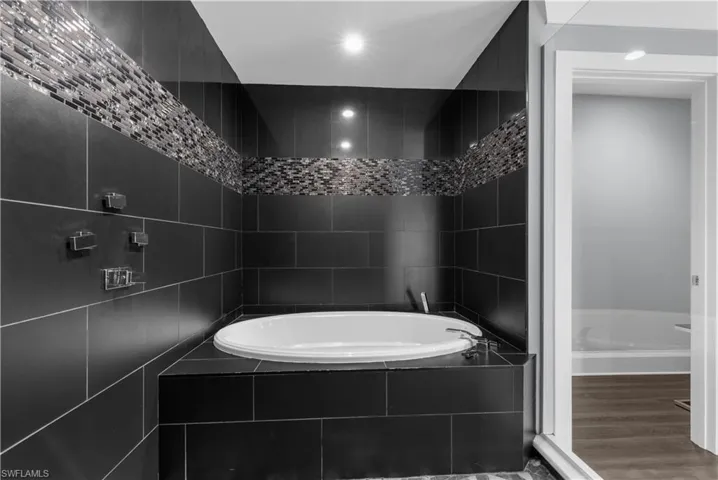Soaking tub set within black subway tile surround, featuring mosaic tile accent band, chrome fixtures, and recessed lighting