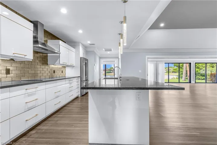 Contemporary kitchen featuring an expansive center island with an undermount sink, pendant lighting, white cabinetry with gold-toned hardware, a stainless steel range hood, and a textured tile backsplash