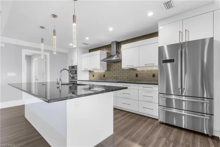 Modern kitchen featuring a large island with a dark countertop, white flat-panel cabinetry, a stainless steel refrigerator, a matching range hood, and wood-finish flooring