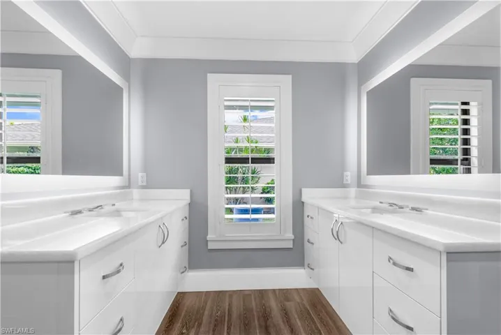 Spacious bathroom featuring dual vanities with white cabinetry and integrated sinks, wood-finish flooring, a central window with plantation shutters, and large wall-mounted mirrors