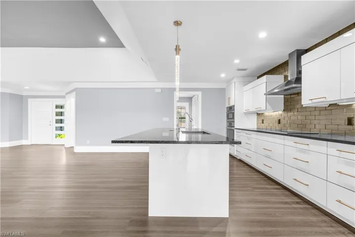 Modern kitchen featuring a spacious island with a dark stone countertop, white high-gloss cabinetry with gold-tone hardware, and a gold-hued subway tile backsplash