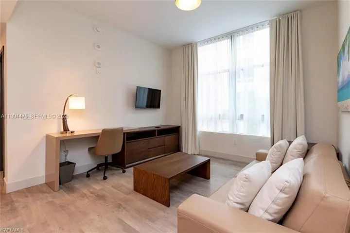 Living area featuring light wood-finish flooring, a large window with sheer and blackout drapes, and a built-in media console