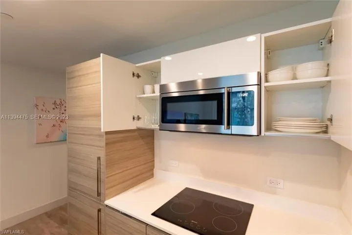 Modern kitchen featuring an integrated stainless steel microwave, white upper cabinetry with interior shelving, light wood-finish tall cabinetry, a sleek black electric cooktop, and light-toned countertops