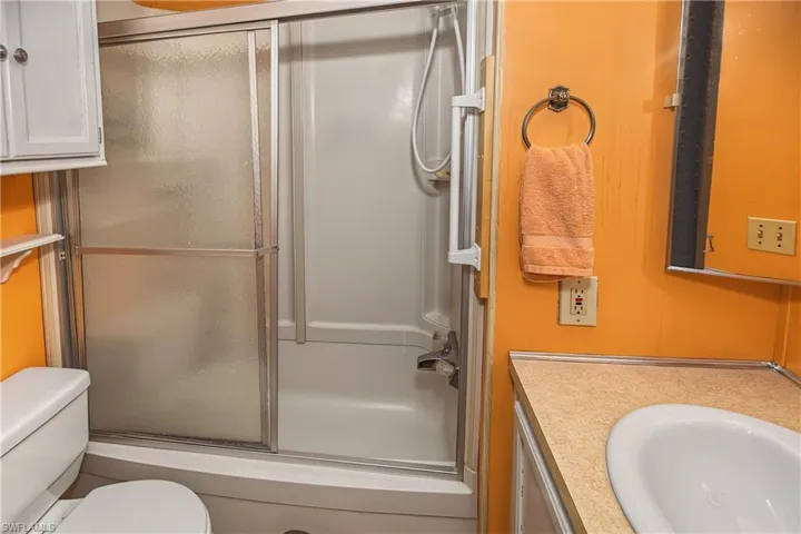 Bathtub with sliding glass enclosure, white toilet, white upper cabinetry, and a vanity with an integrated sink