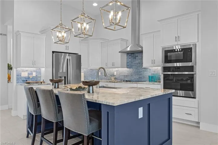 Kitchen with a kitchen island with sink, wall chimney range hood, stainless steel appliances, decorative light fixtures, and white cabinetry