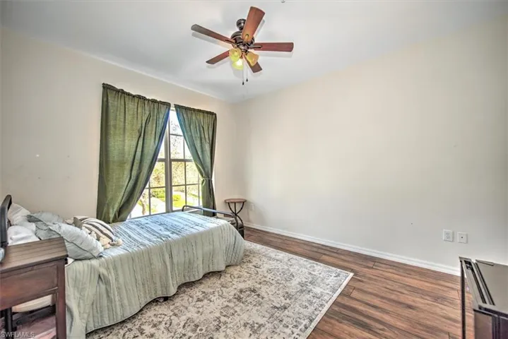 Bedroom featuring a window with green curtains, a ceiling fan with light fixture, wood-finish flooring, and light-toned walls