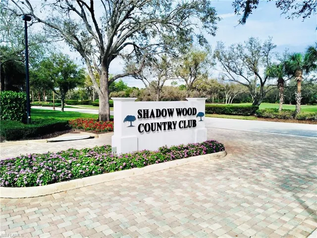 Gated community entrance featuring a prominent monument sign, paver driveway, mature landscaping, and an adjacent golf course