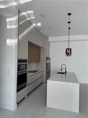 Kitchen with modern cabinets, hanging light fixtures, an island with sink, light stone countertops, and multiple ovens