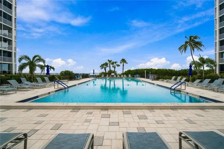 Resort-style swimming pool featuring crystal-clear water, generous sundeck with pavers, dual entry stairs, mature palm landscaping, and direct access to residential towers with lounge seating areas