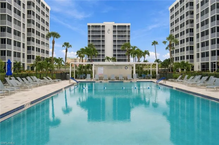 Resort-style swimming pool featuring crystal-clear turquoise water, expansive sun deck with lounge seating, lush tropical landscaping with mature palms, and surrounding multi-story residential towers with modern architectural design