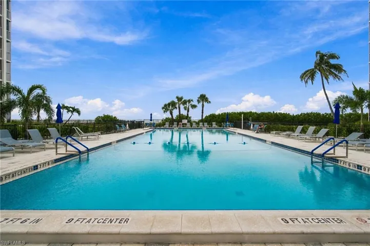 Resort-style saltwater swimming pool featuring crystal-clear turquoise waters, dual entry points with handrails, surrounding lounge deck with integrated seating areas, mature palm tree landscaping, and expansive water views framed by lush tropical grounds