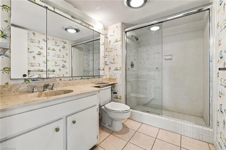 White vanity with a stone countertop and integrated sink, a large mirrored medicine cabinet, a toilet, and a shower with a sliding glass door and white tile surround