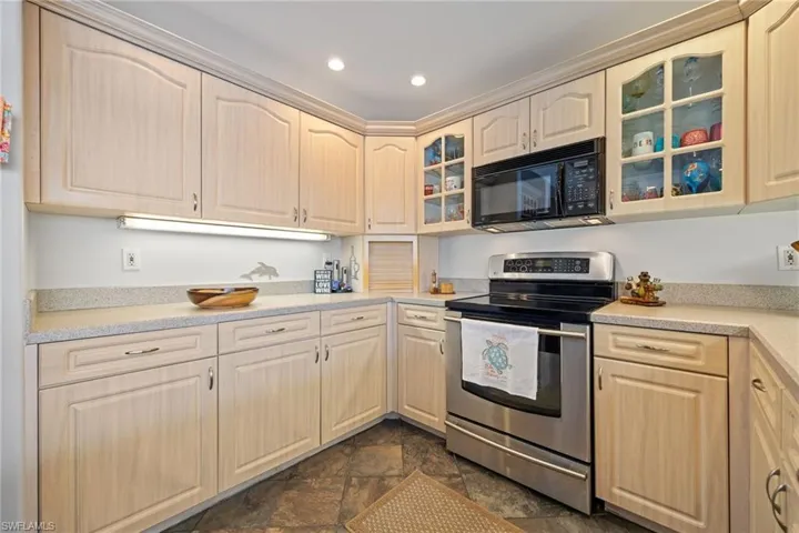 Kitchen with glass insert cabinets, stainless steel range with electric stovetop, black microwave, dark tile flooring, and light wood finish cabinetry